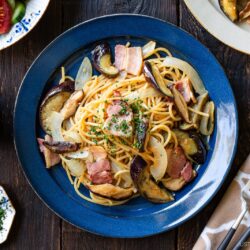 A blue plate of spaghetti topped with bacon, eggplant, onions, and chopped herbs sits on a wooden table, surrounded by salad, cutlery, a pepper grinder, and a small dish of chopped chives.
