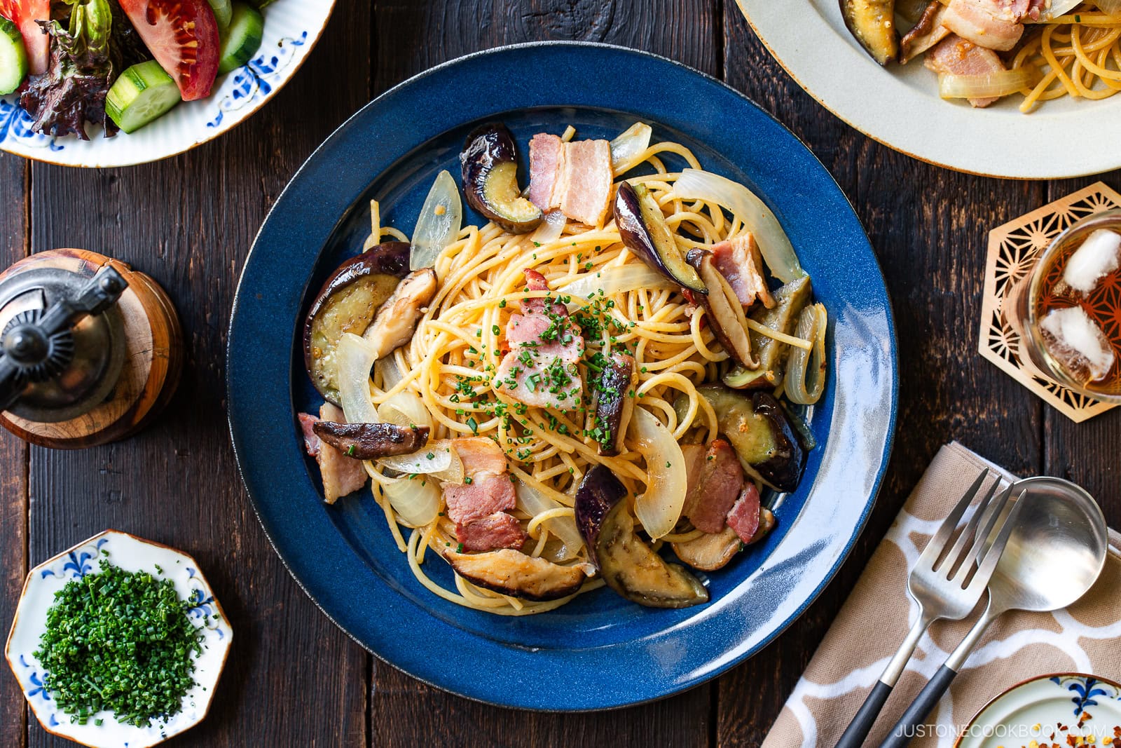 A blue plate of spaghetti topped with bacon, eggplant, onions, and chopped herbs sits on a wooden table, surrounded by salad, cutlery, a pepper grinder, and a small dish of chopped chives.