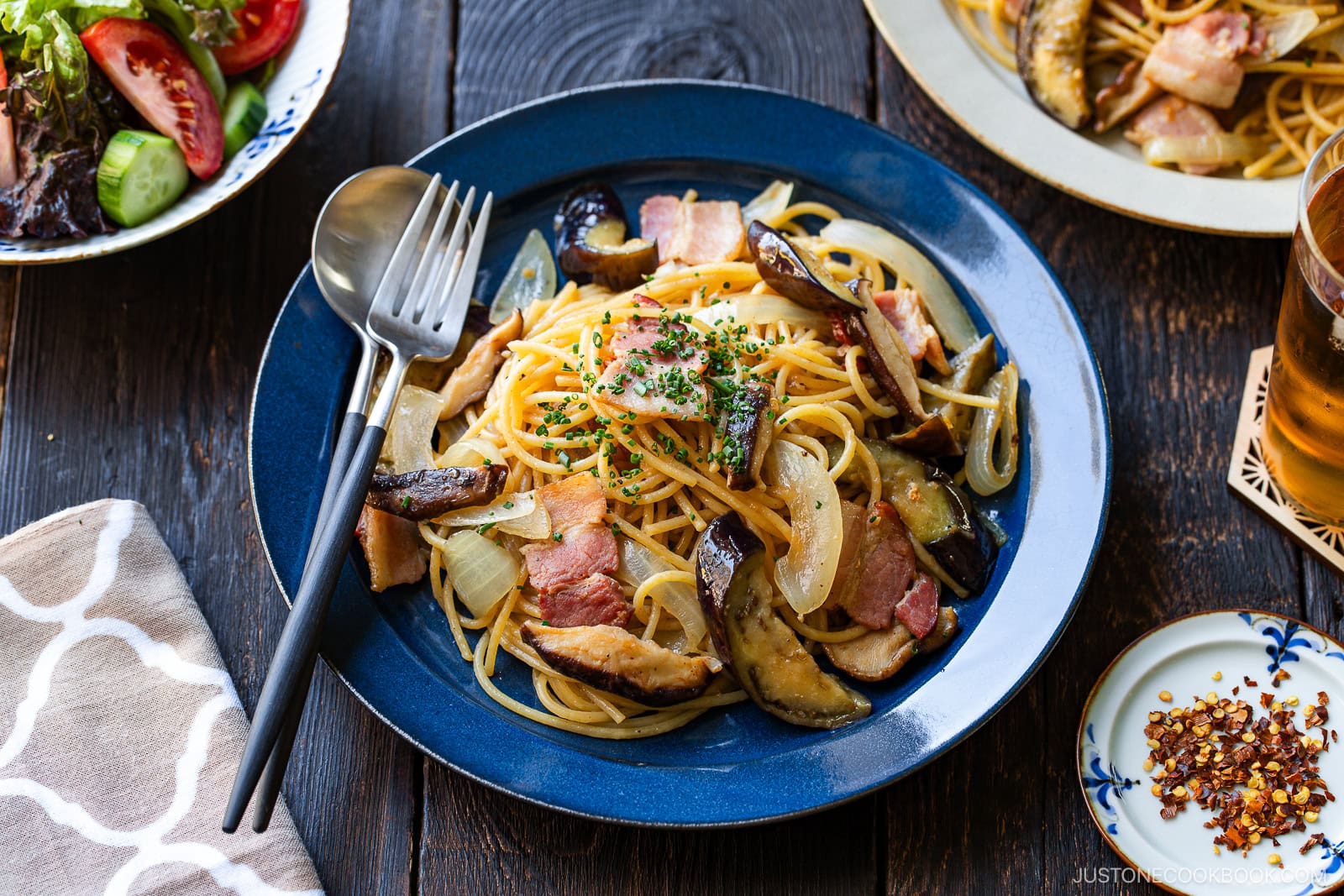 A plate of spaghetti topped with sautéed eggplant, onions, bacon, and chopped herbs, served with a fork and spoon. A side salad, a small dish of red pepper flakes, and a glass of drink are nearby on a rustic wooden table.