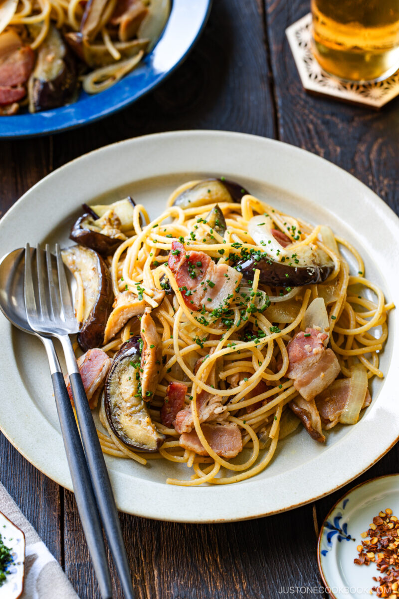 A plate of spaghetti topped with bacon, eggplant, onions, and chopped herbs, served with a fork and spoon on the side. A glass of beer is in the background on a wooden table.
