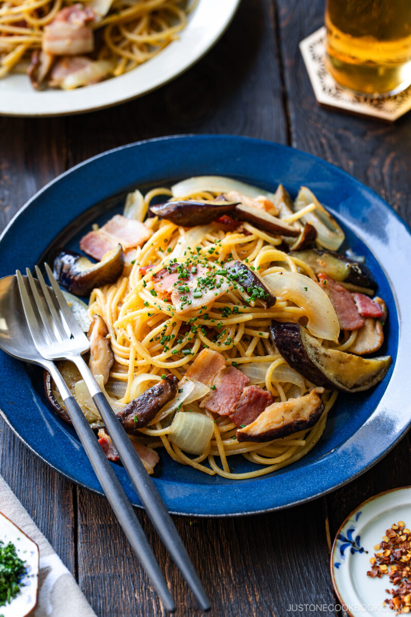 A blue plate of spaghetti topped with bacon, sautéed onions, and mushrooms, garnished with chopped herbs. A fork and spoon rest on the plate; a glass of beer and another dish are in the background on a wooden table.