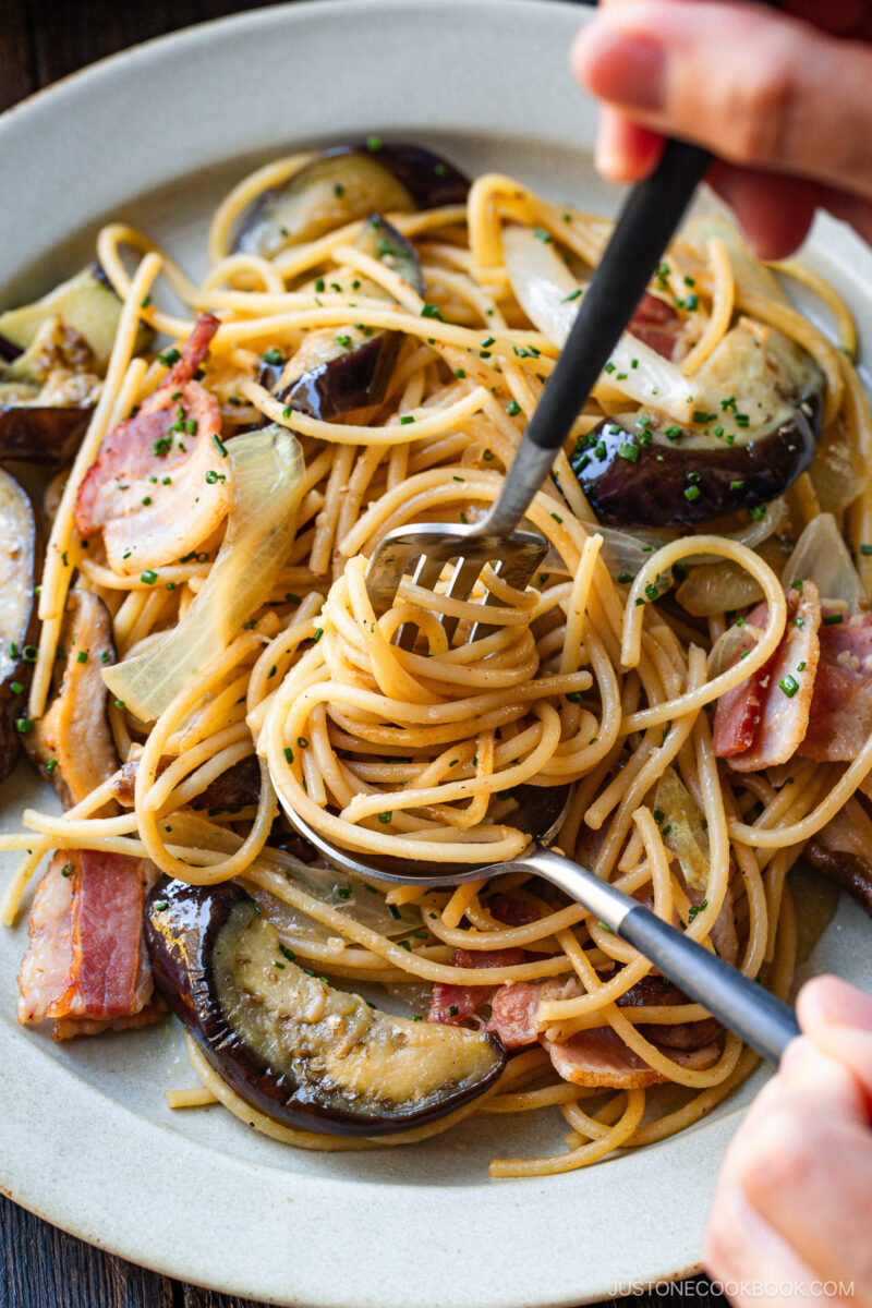 A close-up of a plate of spaghetti with slices of eggplant, bacon, and onion, garnished with chopped herbs. Two hands are twirling the pasta with a fork and spoon.