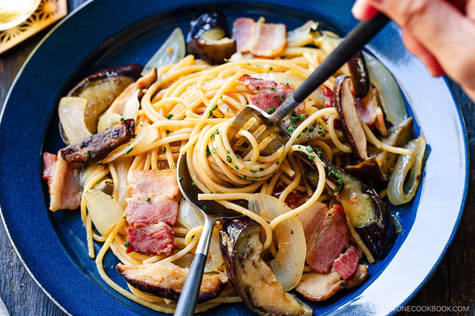 A close-up of a plate of pasta with slices of bacon, sautéed onions, eggplant, and herbs. A hand uses utensils to twirl the pasta on a dark blue plate.
