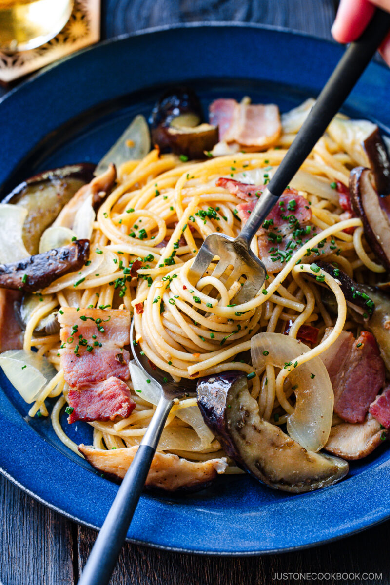 A blue plate of spaghetti with sautéed onions, bacon, mushrooms, and herbs. A fork and spoon are twirling the pasta, and a glass of drink is partially visible in the background.