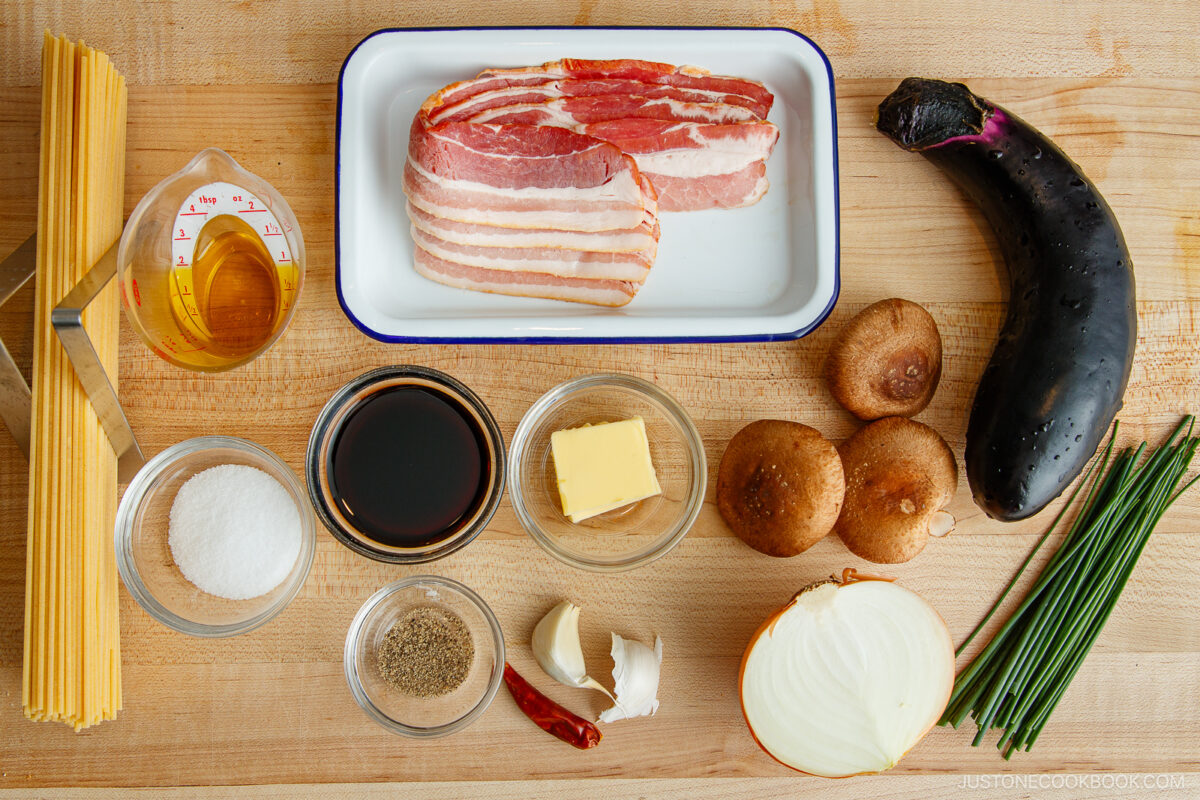 A wooden surface displays uncooked spaghetti, bacon slices, an eggplant, shiitake mushrooms, chives, a sliced onion, butter, soy sauce, sugar, black pepper, cooking oil in a measuring cup, garlic cloves, and a dried chili pepper.