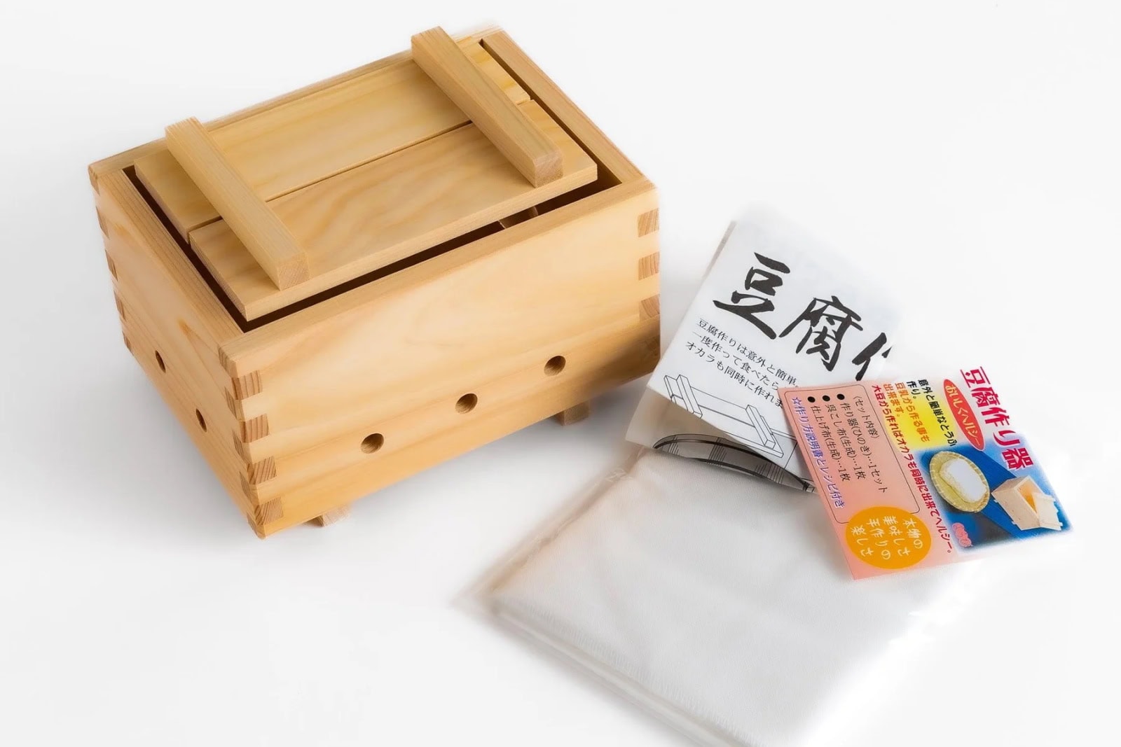 A wooden tofu press box with a lid is shown next to a folded white cloth and packaged tofu-making supplies with Japanese text.