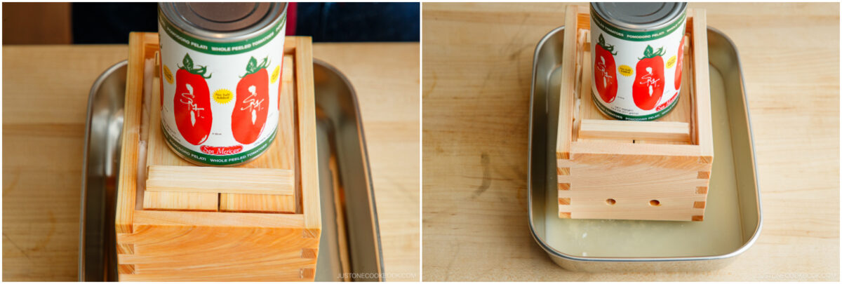 A metal can is placed on top of a wooden tofu press, which sits in a shallow metal tray containing liquid, on a wooden surface.
