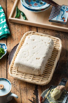 A block of tofu sits on a rectangular woven bamboo tray, surrounded by Japanese-style dishes, a green napkin with white spots, and a wooden table surface.