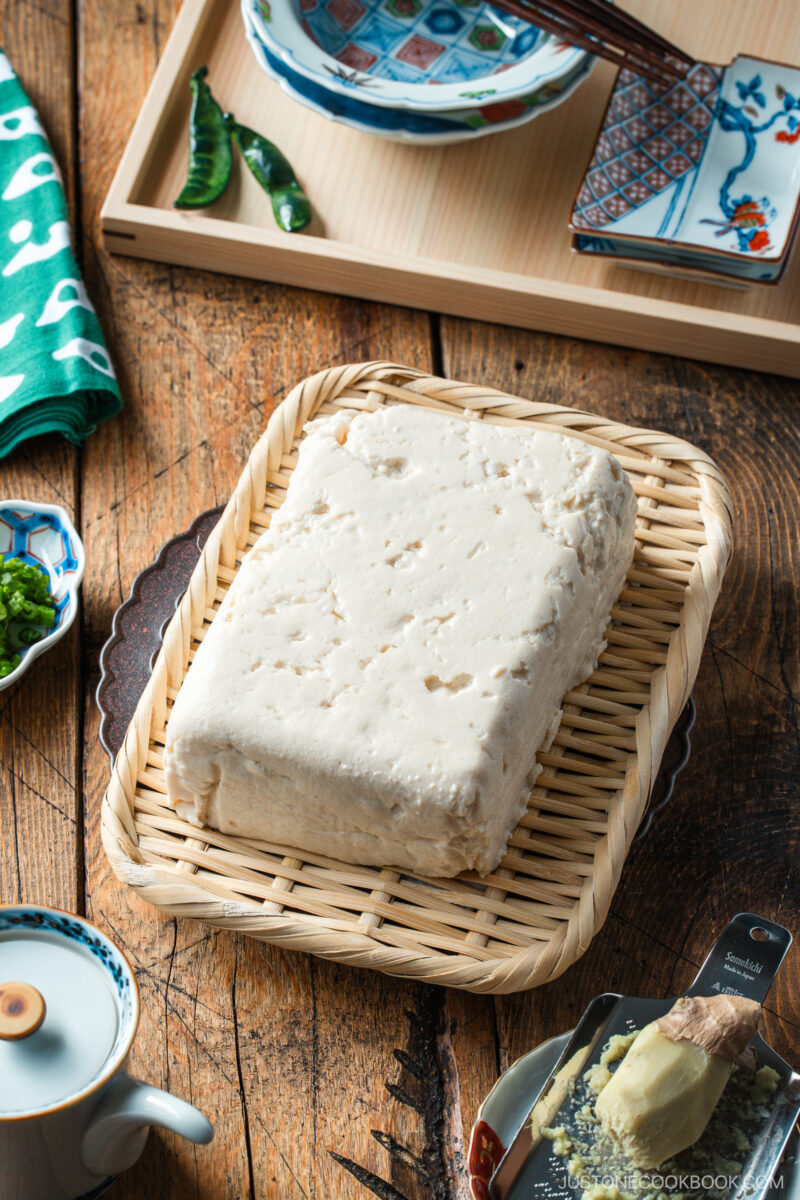 A block of tofu sits on a rectangular woven bamboo tray, surrounded by Japanese-style dishes, a green napkin with white spots, and a wooden table surface.