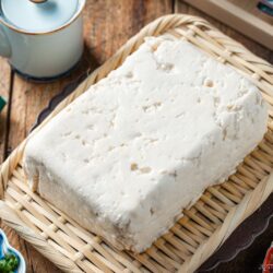 A block of fresh tofu rests on a woven bamboo tray, surrounded by various small dishes and utensils on a wooden table.