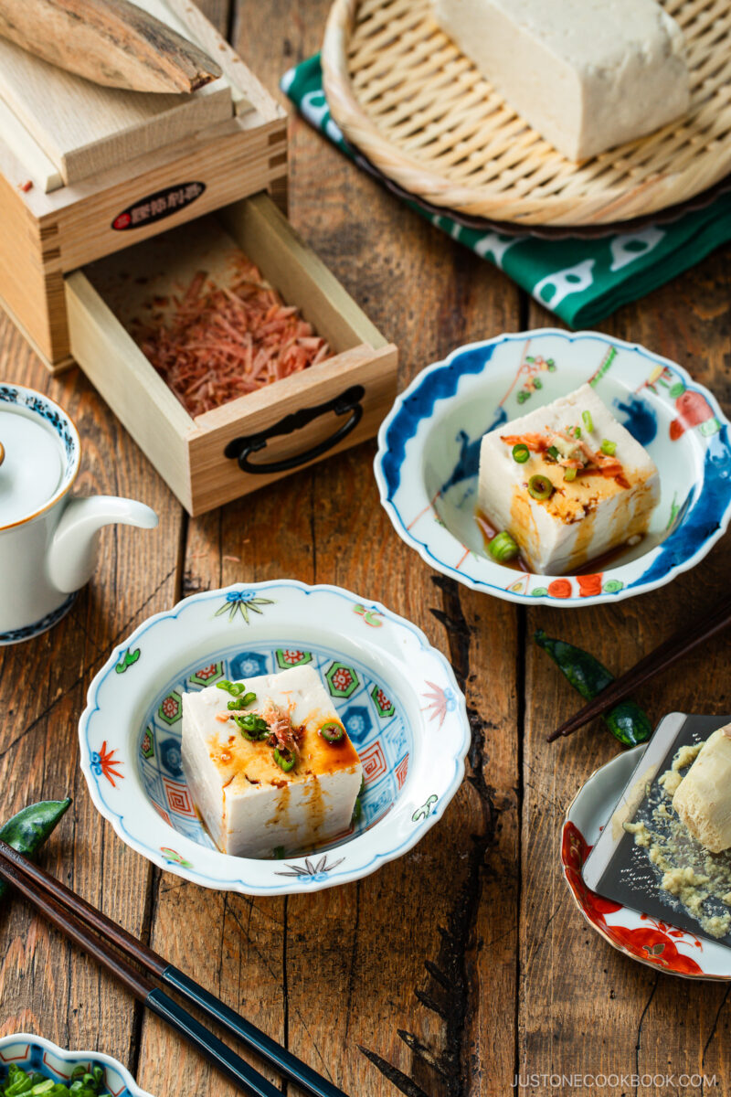 Two bowls of silken tofu topped with green onions and sauce sit on a wooden table, accompanied by chopsticks, a teapot, a box of dried bonito flakes, and a basket holding a block of tofu.