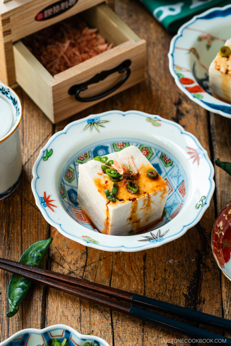 A bowl with a block of silken tofu topped with sliced green onions, bonito flakes, and a drizzle of soy sauce, on a wooden table with chopsticks and other dishes nearby.