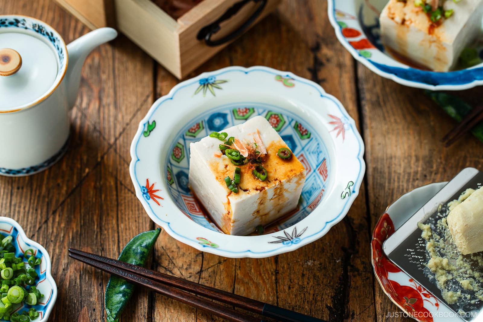 A bowl with a block of tofu topped with chopped green onions and sauce sits on a wooden table, surrounded by chopsticks, a teapot, and other small dishes with condiments and tofu.
