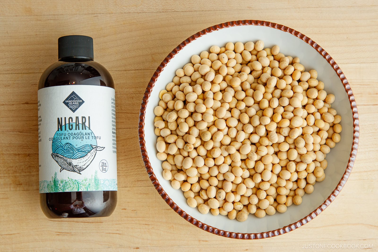 A bottle of Nigari tofu coagulant sits next to a bowl filled with dried soybeans on a light wooden surface.