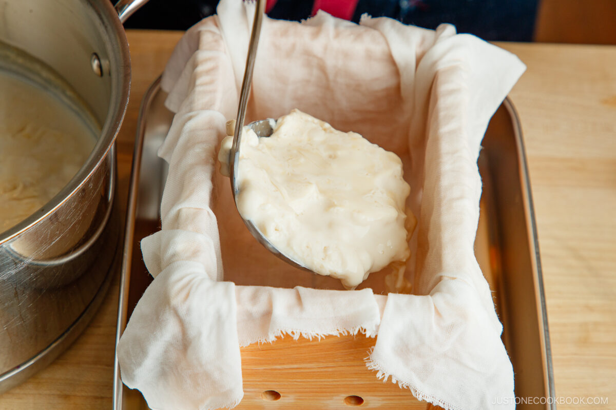 A ladle pours fresh curds onto a cheesecloth-lined mold, set on a tray, as part of the homemade tofu-making process. A pot with more curds is visible to the side.