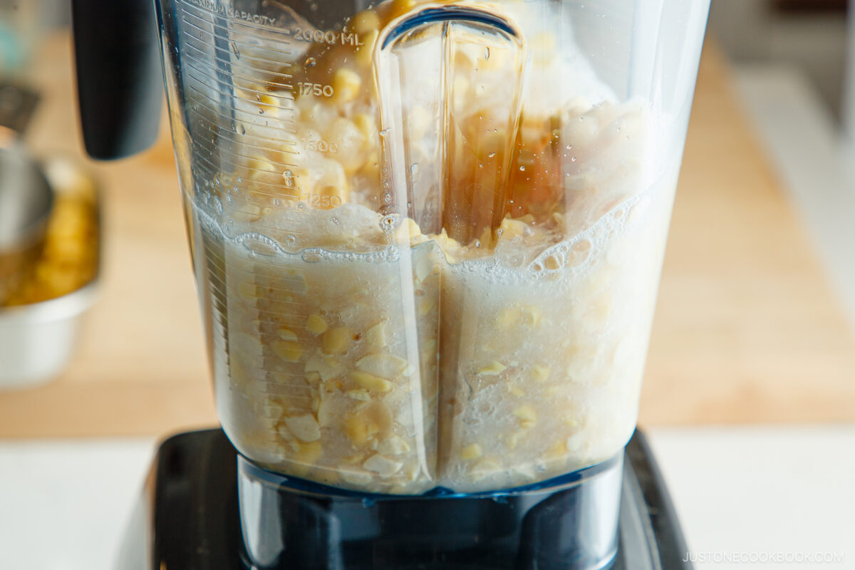 Close-up of a blender containing corn kernels and liquid being blended, with froth forming at the top. The blender is on a countertop, and a kitchen background is visible.