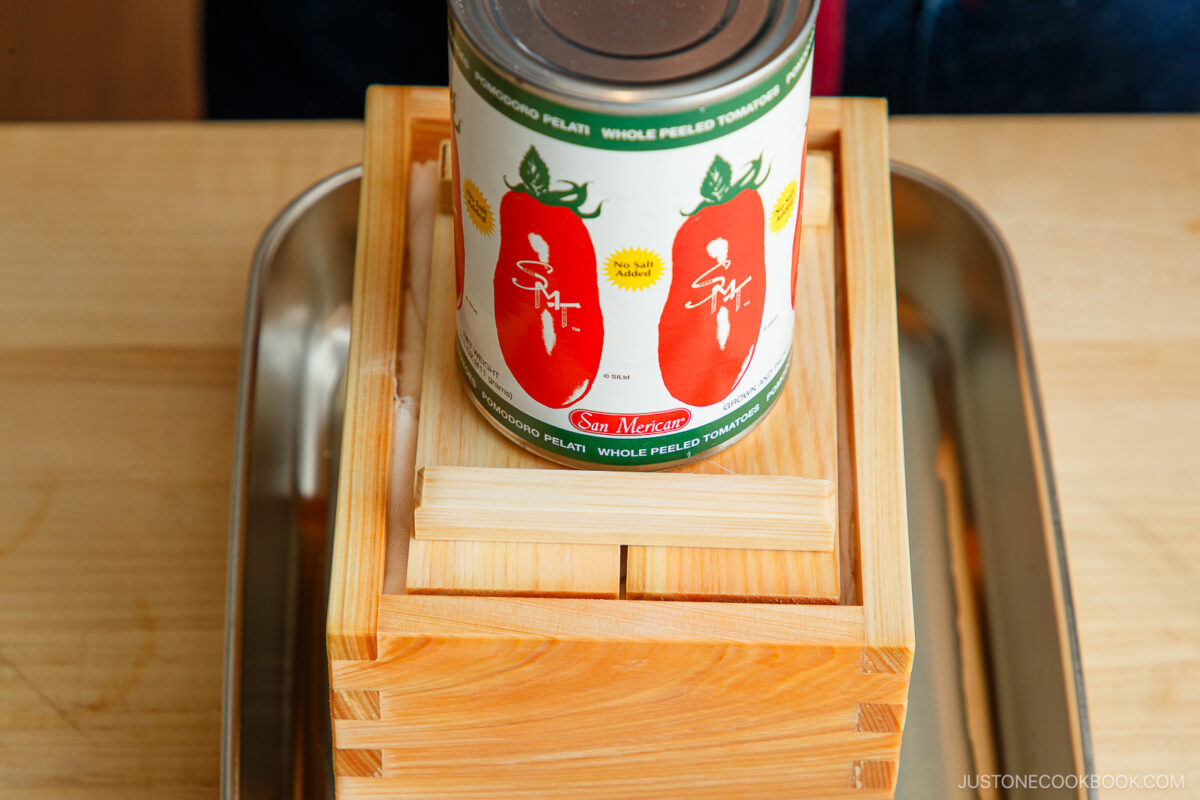 A can of San Marzano whole peeled tomatoes sits on top of a wooden tofu press, which is placed on a metal tray on a wooden surface.