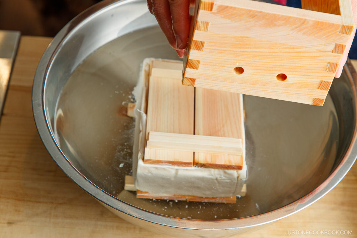 A person presses tofu with a wooden box mold over a metal bowl, draining excess water as part of the tofu-making process.