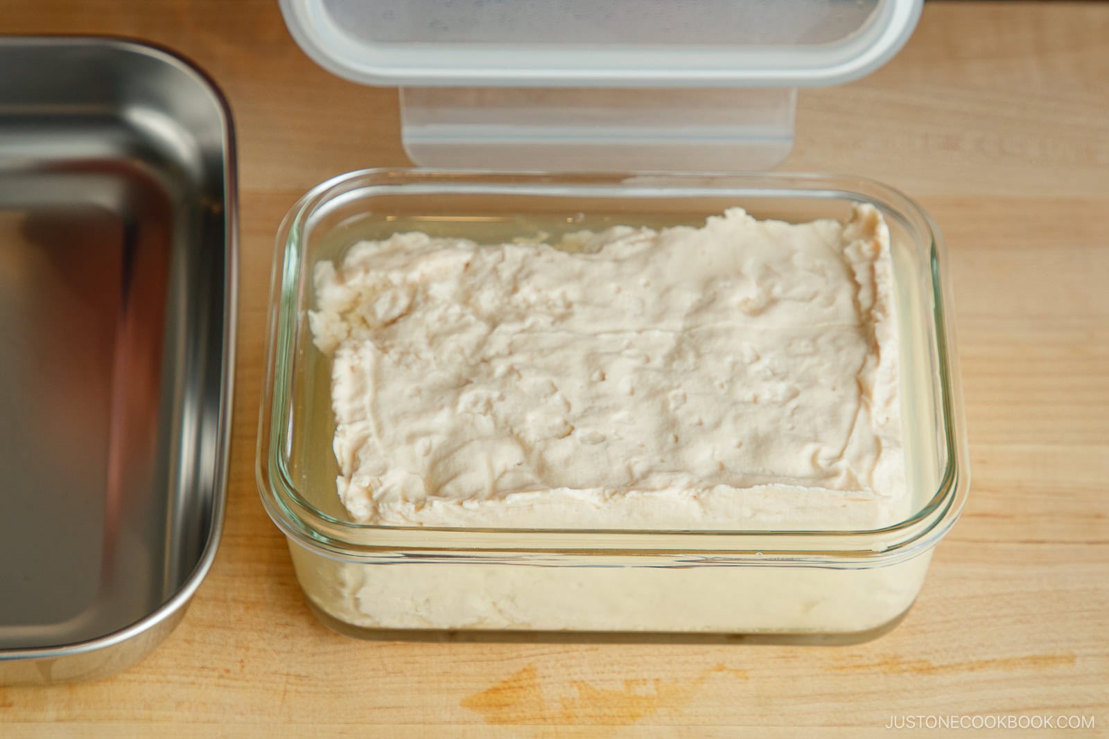 A rectangular block of tofu sits in a clear glass container with a lid, placed on a wooden surface. Part of a metal tray is visible to the side.