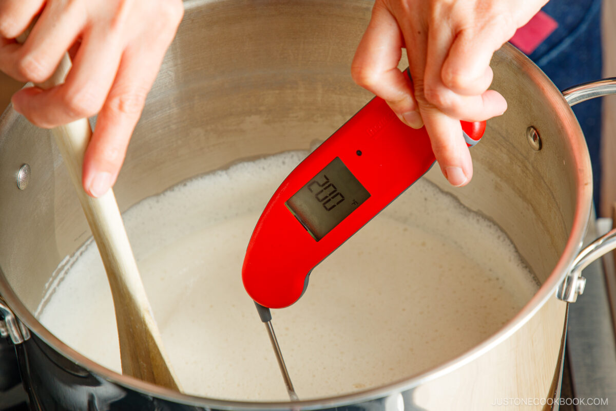 A person holds a red digital thermometer reading 200°F above a pot of simmering milk while stirring with a wooden spoon.