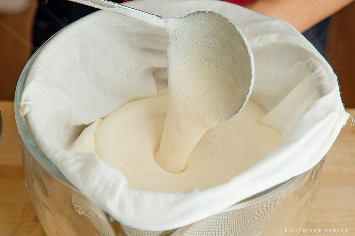 A ladle pours foamy soy milk through a cloth-lined strainer set over a glass bowl, demonstrating the process of filtering homemade soy milk.