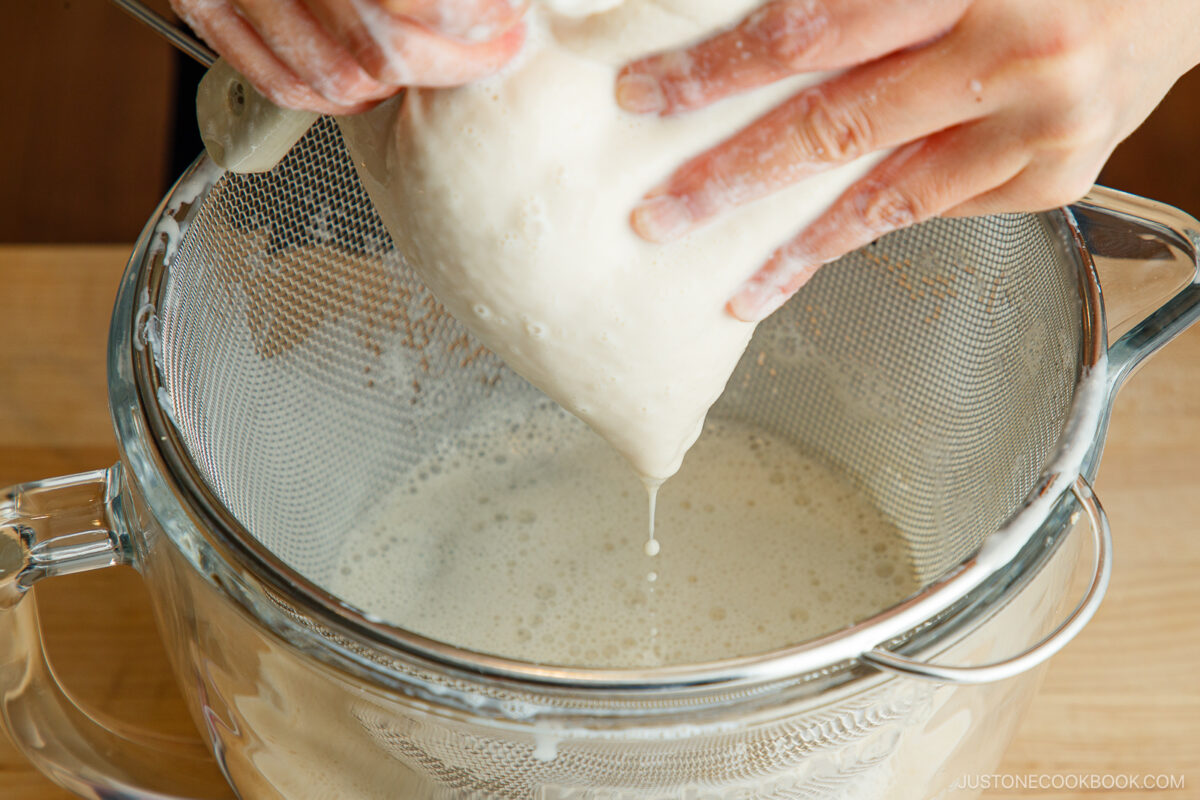 A person squeezes a nut milk bag filled with blended soybeans over a fine mesh strainer, filtering homemade soy milk into a glass bowl below.