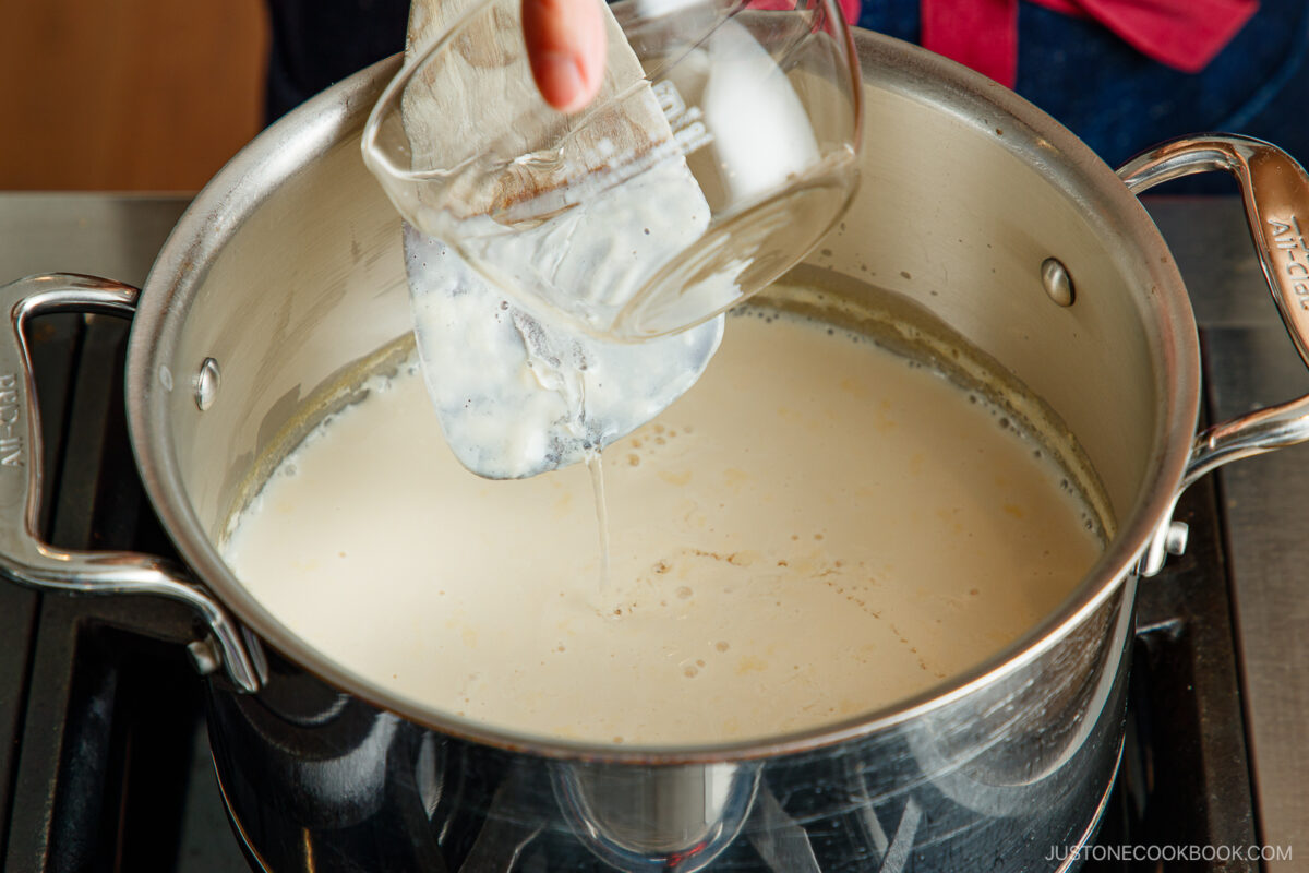 A hand pours a liquid from a glass measuring cup into a pot of creamy mixture on a stovetop.