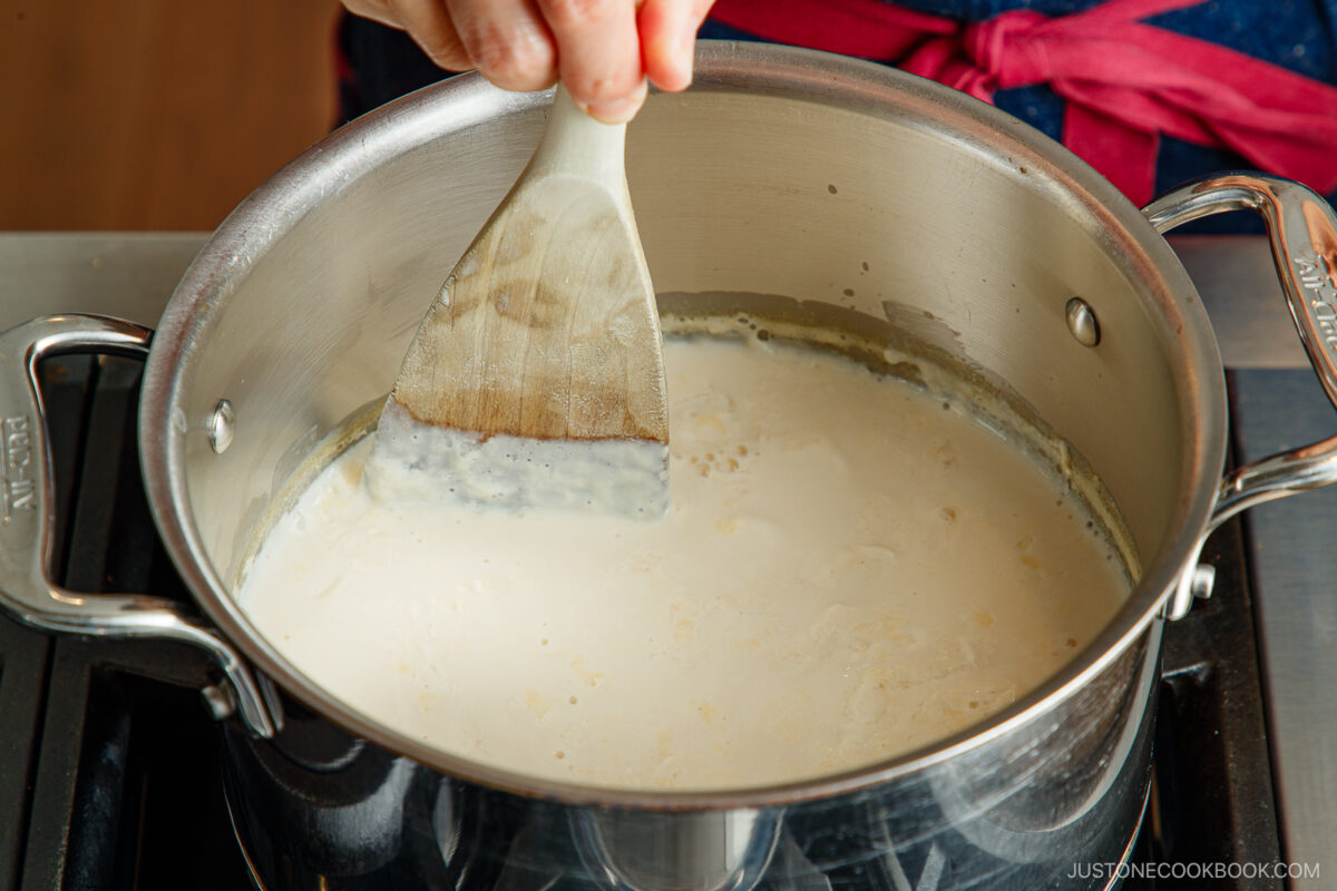 A person stirs a creamy white mixture in a stainless steel pot on a stovetop using a wooden spatula. The person is wearing a blue apron with a red tie.