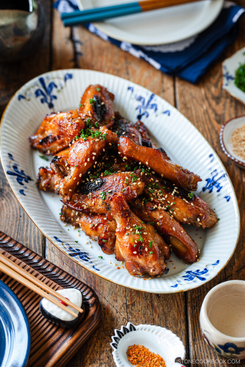 A white oval dish with blue patterns holds glazed, baked chicken wings sprinkled with sesame seeds and chopped herbs, placed on a rustic wooden table set with chopsticks, plates, and small bowls.