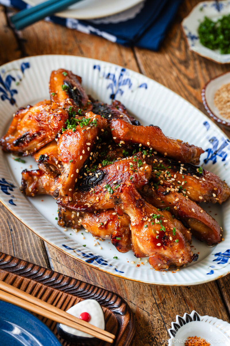 A plate of glazed grilled chicken wings garnished with chopped green herbs and sesame seeds, served on a decorative white and blue dish on a wooden table. Chopsticks and small bowls are visible nearby.