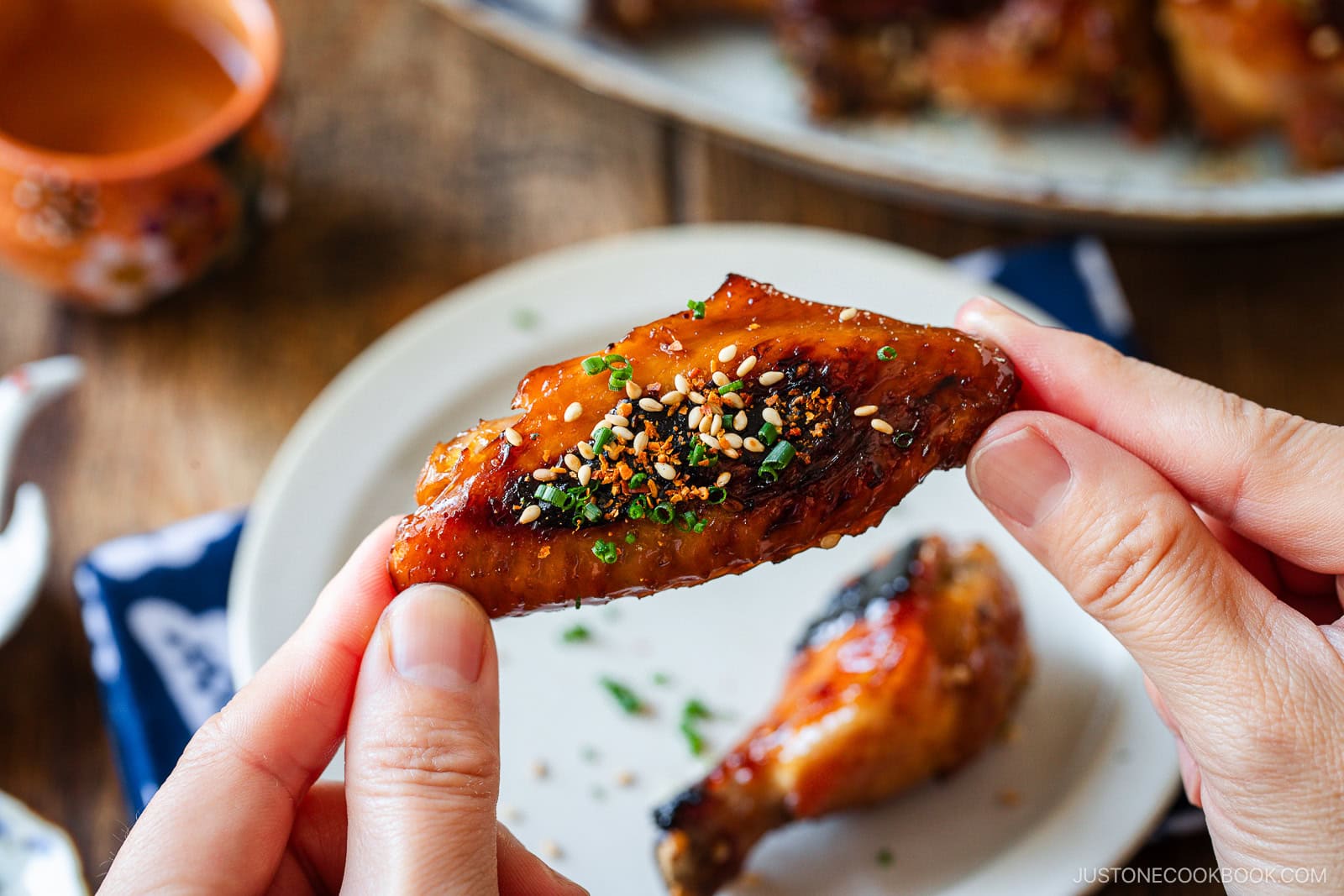 A close-up of hands holding a glazed chicken wing garnished with sesame seeds and herbs, with a plate of more chicken and a cup in the background on a wooden table.