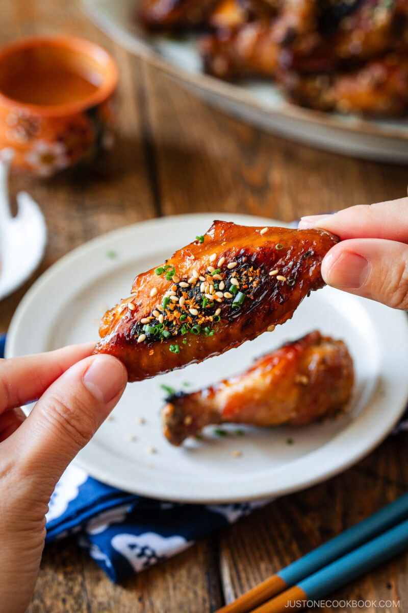 A hand holds a glazed chicken wing topped with sesame seeds and chopped green onions; more chicken wings sit on a white plate in the background, with chopsticks and a cup nearby on a wooden table.