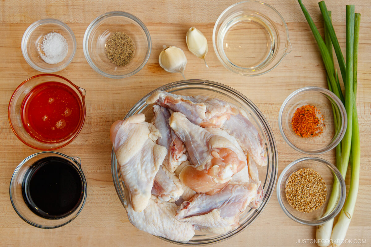 Raw chicken wings in a glass bowl surrounded by small bowls of soy sauce, hot sauce, oil, vinegar, garlic cloves, salt, pepper, chili flakes, sesame seeds, and two stalks of green onion on a wooden surface.
