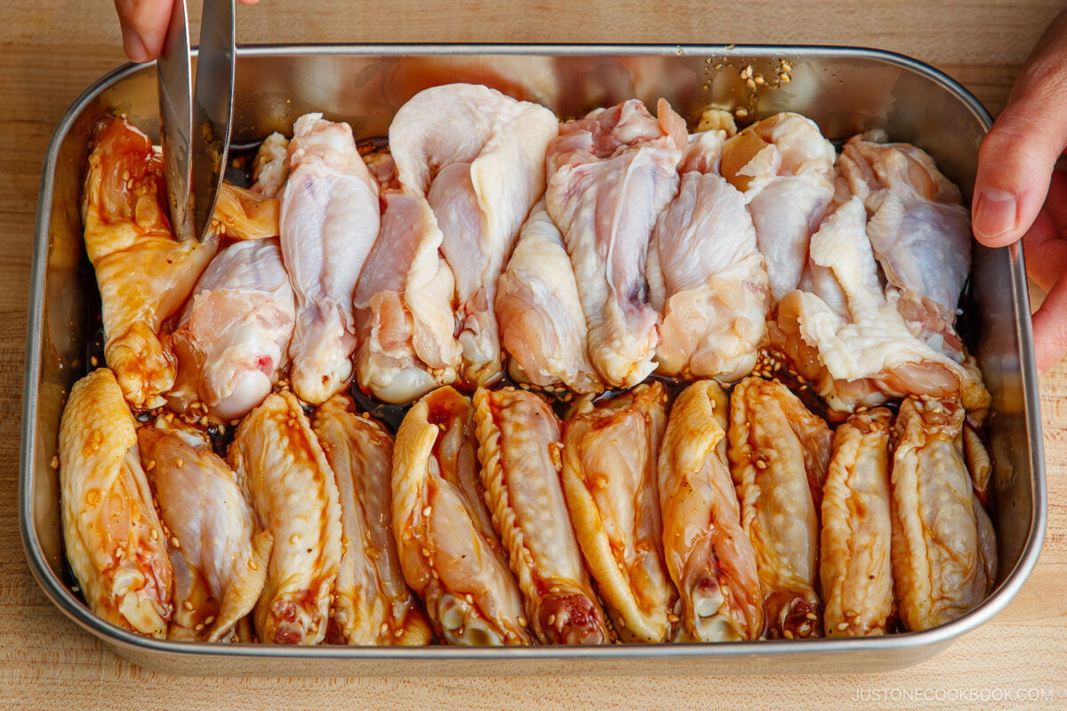 A metal tray filled with raw chicken wings marinated in sauce; a hand uses tongs to arrange the pieces on a wooden surface.