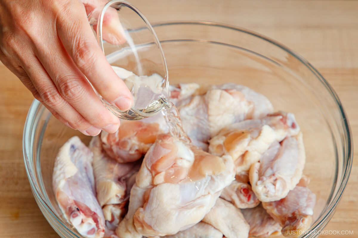 A hand pours liquid from a small glass cup over raw chicken wings in a clear glass bowl, preparing them on a wooden surface.