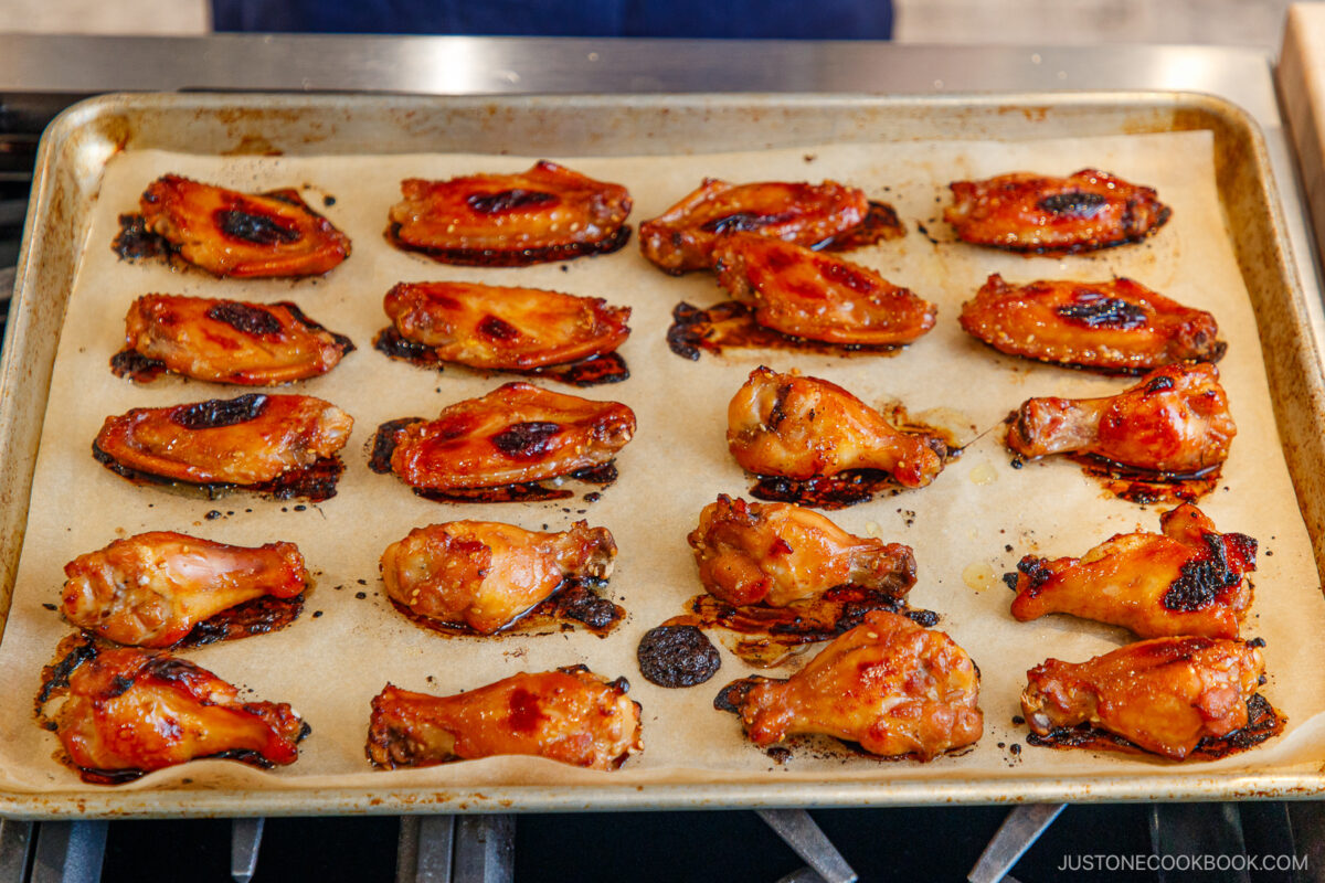 A baking sheet lined with parchment paper holds rows of glazed chicken wings, some with caramelized and slightly charred edges, fresh out of the oven.