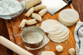 A wooden board with round dumpling wrappers, dough balls, a rolling pin, a metal cutter, cut dough pieces, and a bowl of flour, showing the process of making dumpling wrappers.