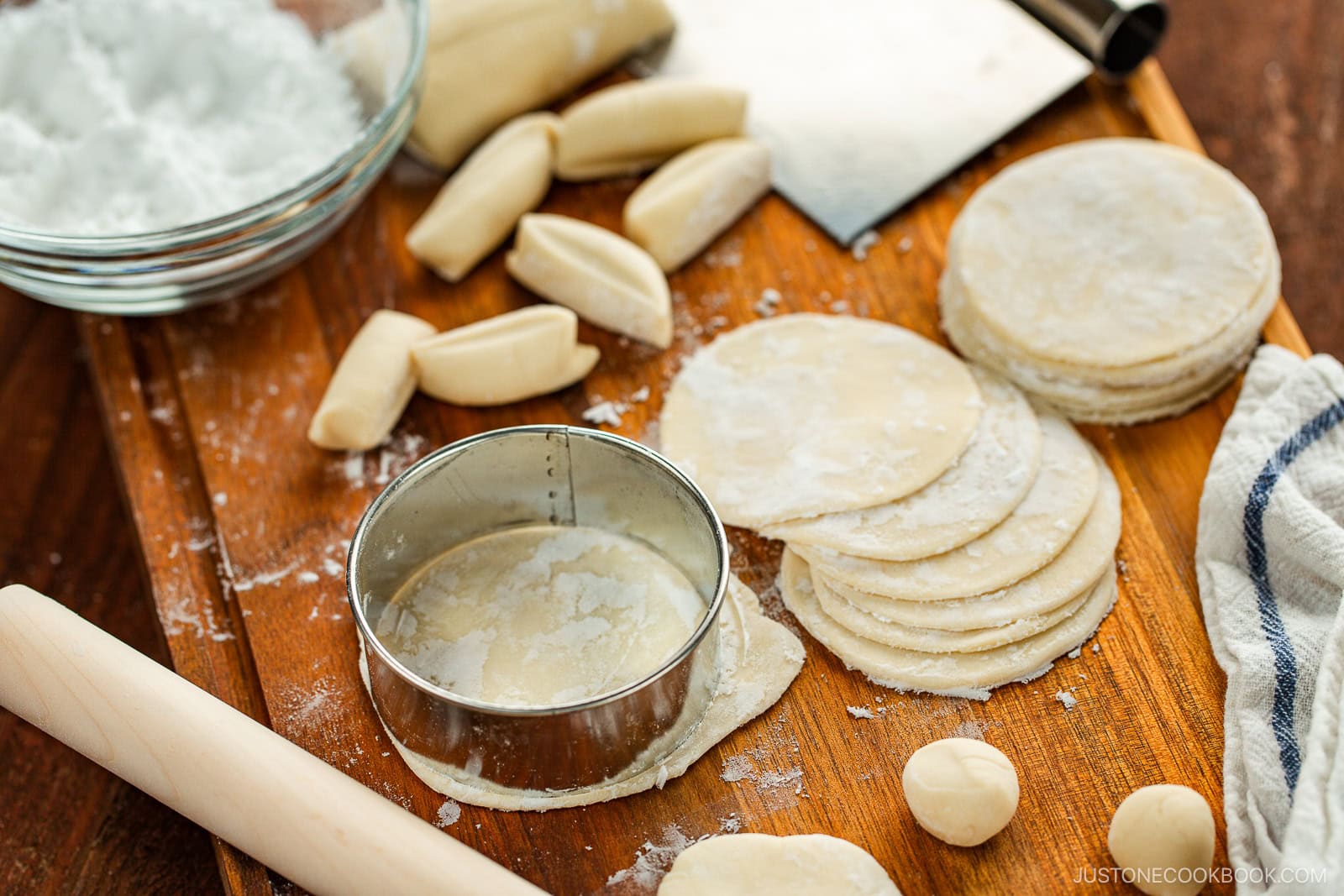 A wooden board with round dumpling wrappers, dough balls, a rolling pin, a metal cutter, cut dough pieces, and a bowl of flour, showing the process of making dumpling wrappers.