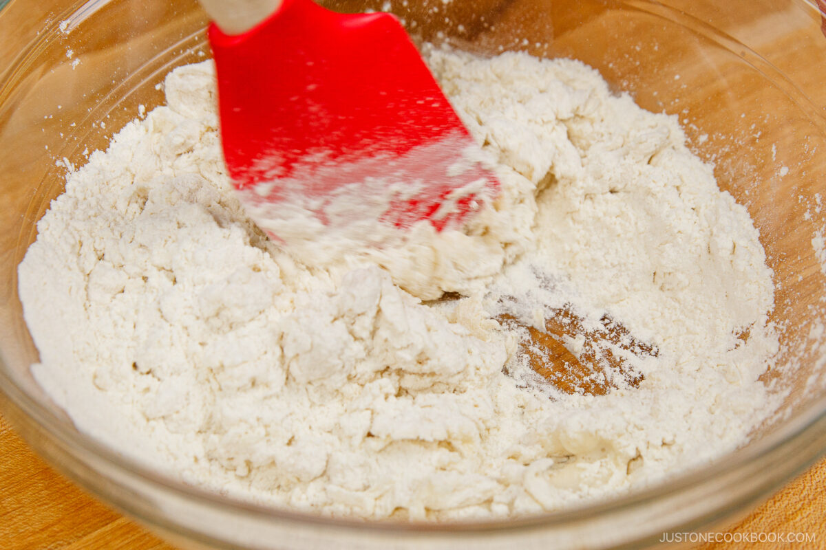 A red spatula mixes flour and other dry ingredients in a clear glass bowl on a wooden surface.