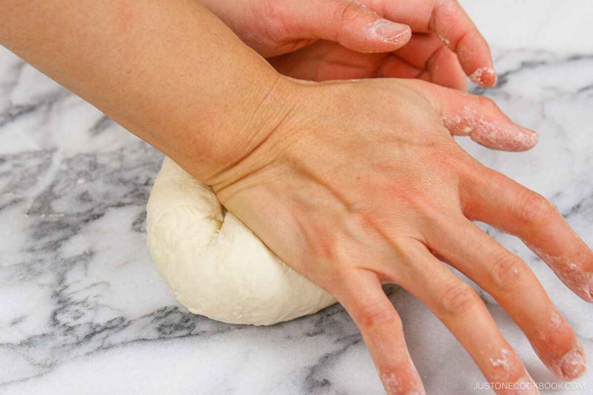A close-up of hands kneading a ball of dough on a marble surface, with a light dusting of flour visible.