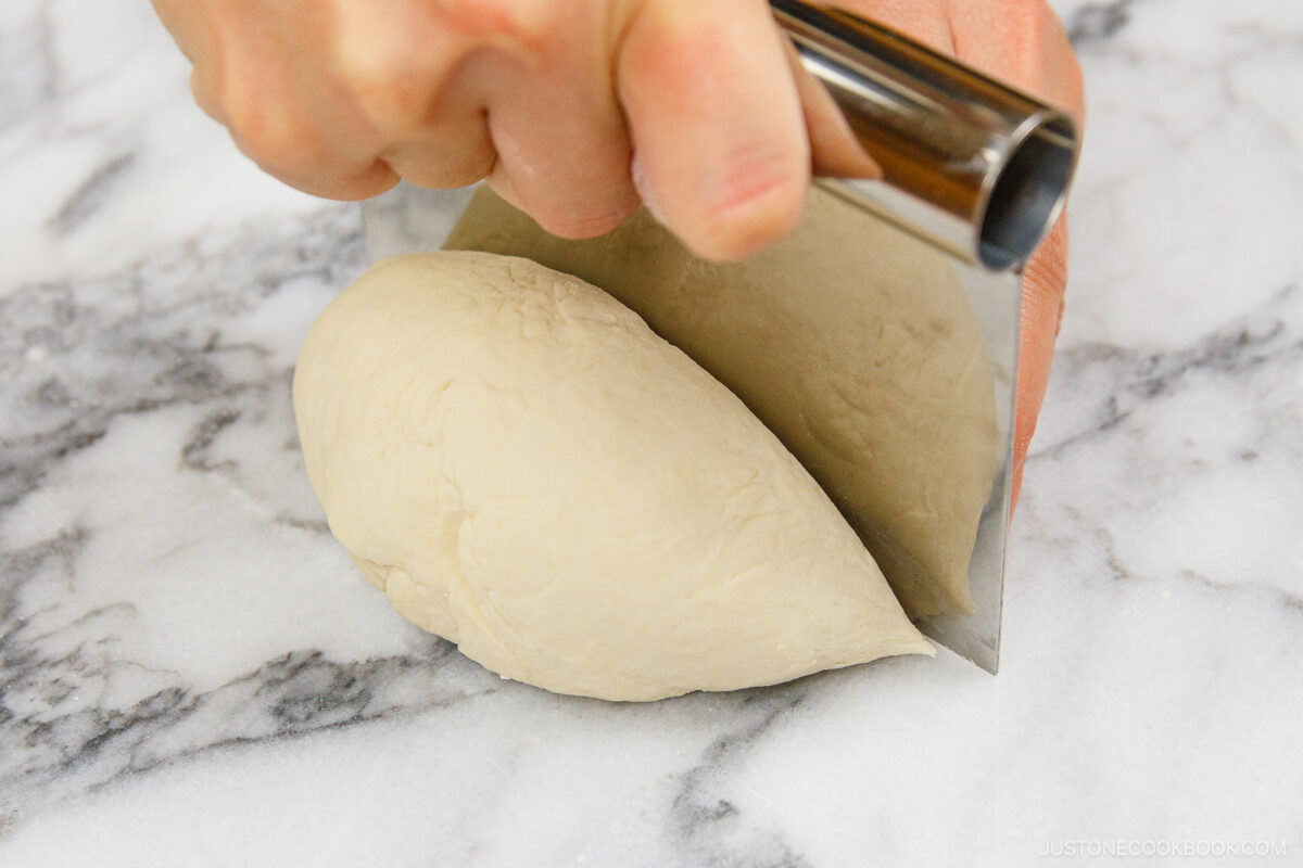 A hand holds a metal bench scraper, cutting a ball of dough on a white marble surface.