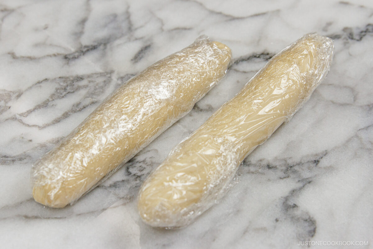 Two logs of dough wrapped in plastic wrap are placed on a marble surface, ready to be chilled.