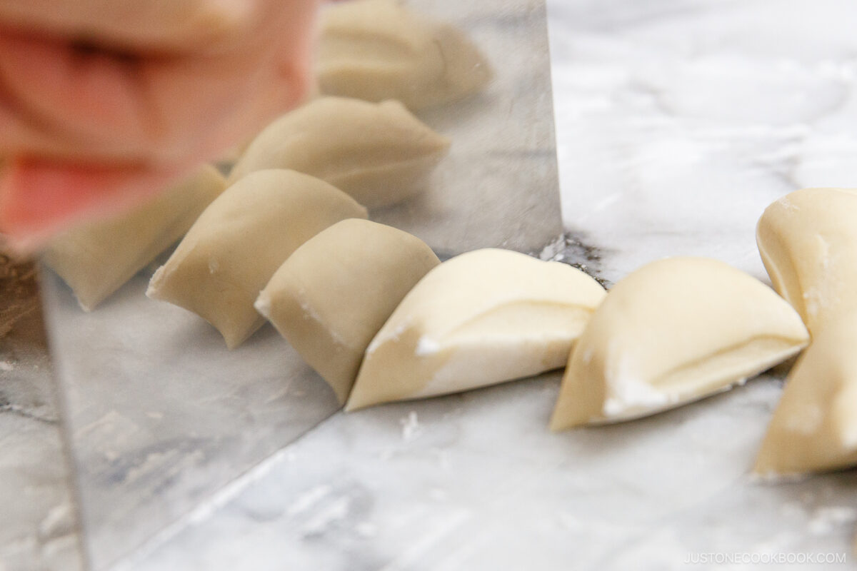 A close-up of a hand using a dough scraper to cut a log of dough into small, even pieces on a floured marble surface.