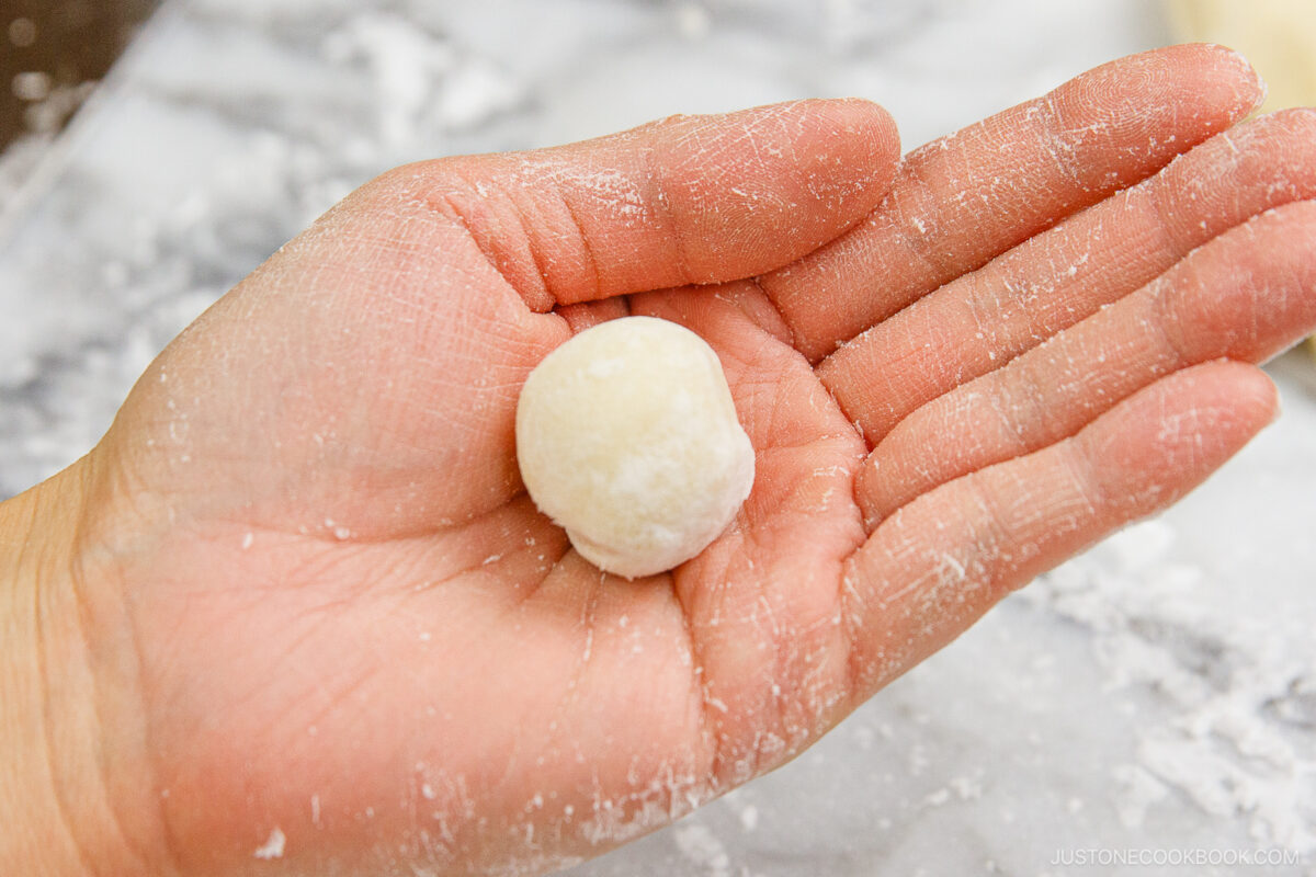 A close-up of a hand holding a small, round ball of dough dusted with flour, with a floured surface in the background.