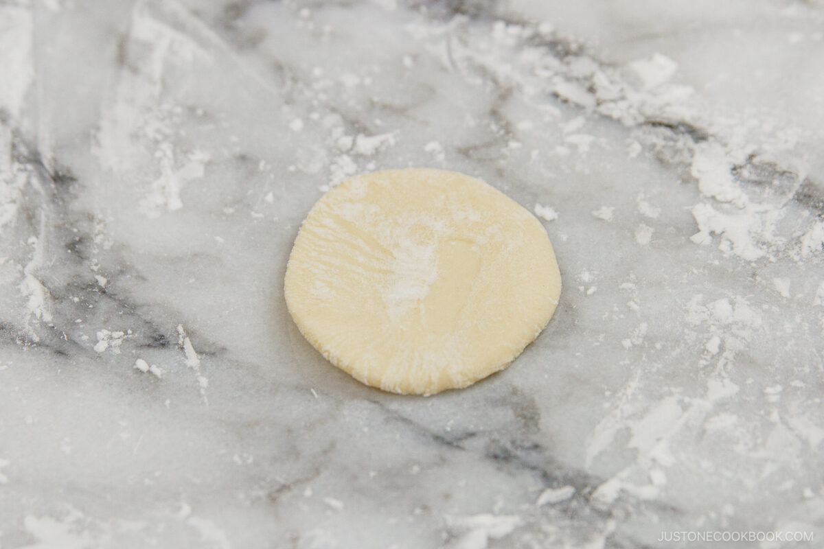 A round, flat disc of dough dusted with flour rests on a lightly floured marble surface.
