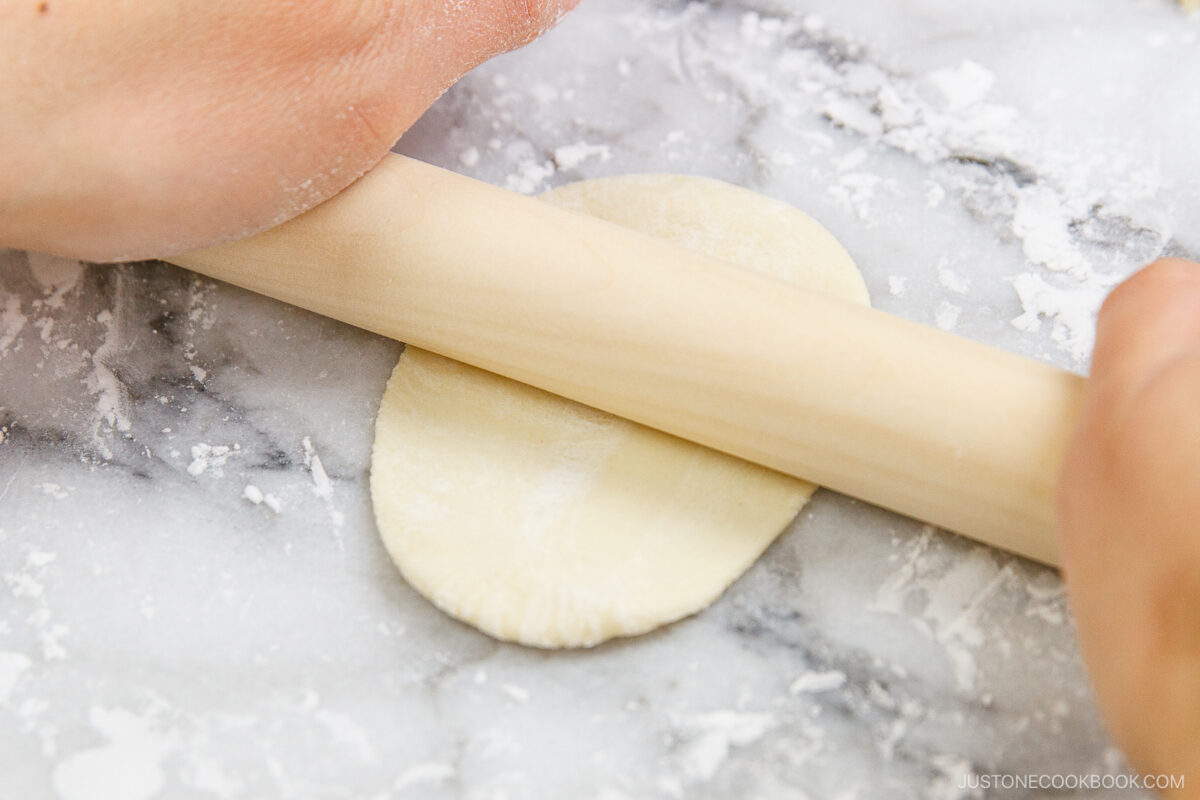 Close-up of hands using a rolling pin to flatten dough on a floured marble surface.