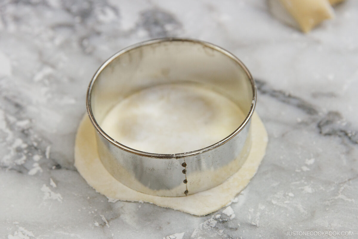 A round metal cookie cutter sits on top of a flat circular sheet of dough on a marble surface. Partially rolled dough is visible in the background.