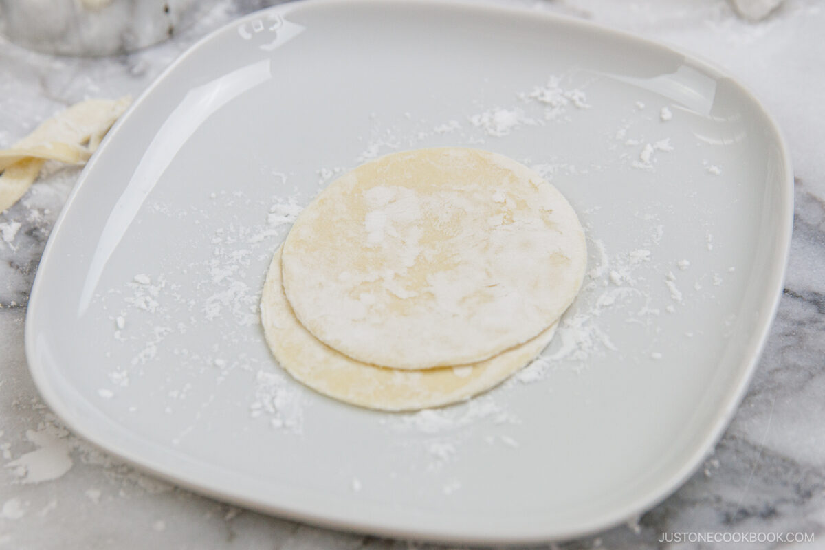 Two round, uncooked dumpling wrappers dusted with flour sit on a white plate, with a marble surface and some flour in the background.