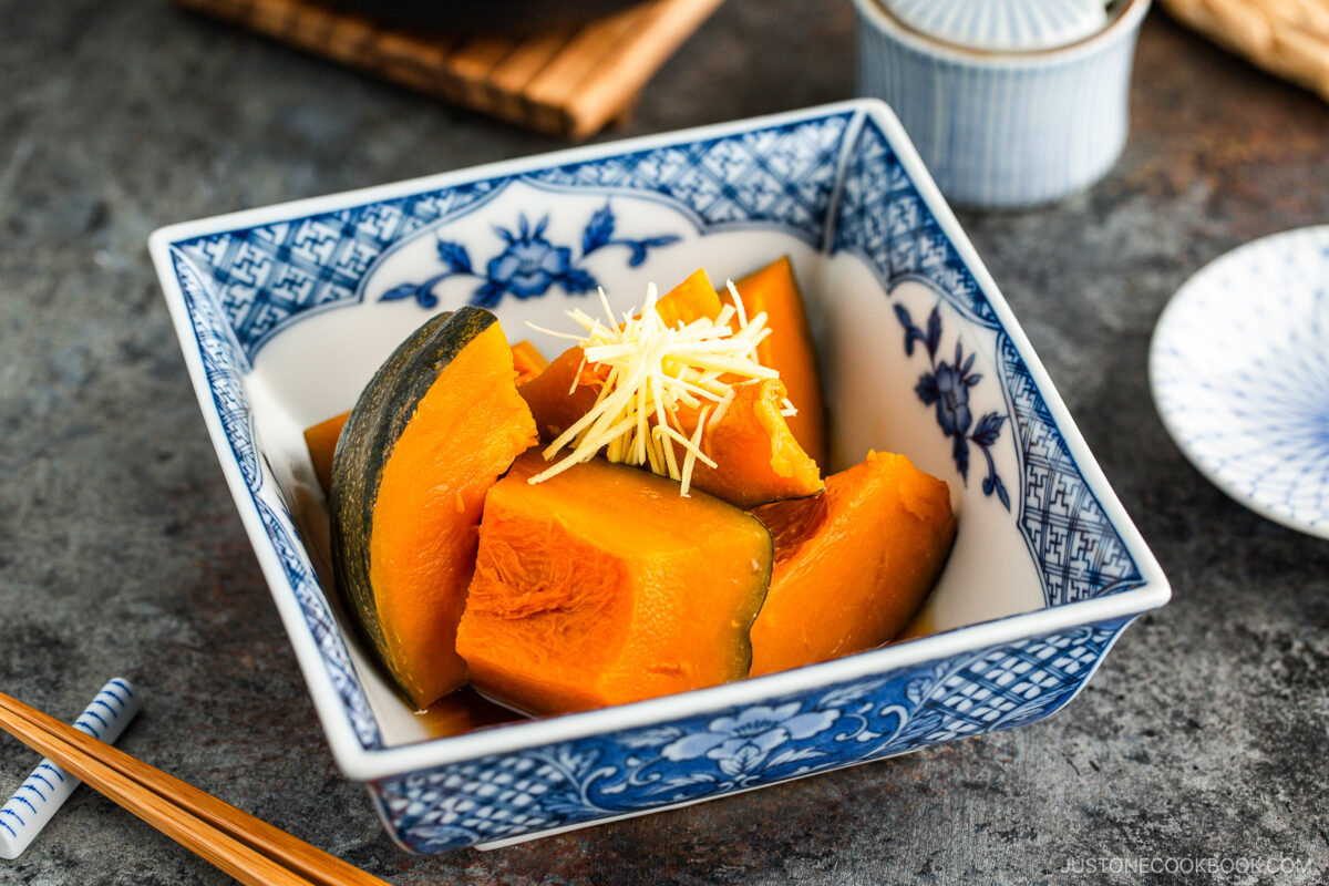 A decorative blue-and-white bowl filled with simmered kabocha squash pieces, garnished with thinly sliced ginger. Chopsticks and small dishes are placed beside the bowl on a dark surface.