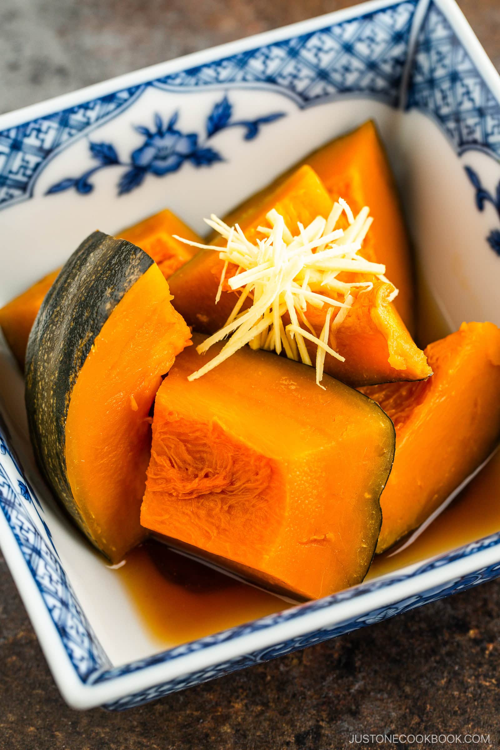 Four pieces of simmered kabocha squash in a blue and white patterned dish, topped with finely shredded ginger. The squash is orange with green skin and is served in a light sauce.